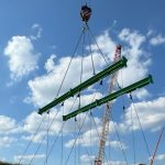 A crane hoists two green steel beams into the air at a construction site, with two red and white cranes and a blue sky with clouds in the background, seamlessly blending technology with precision like an auto draft.