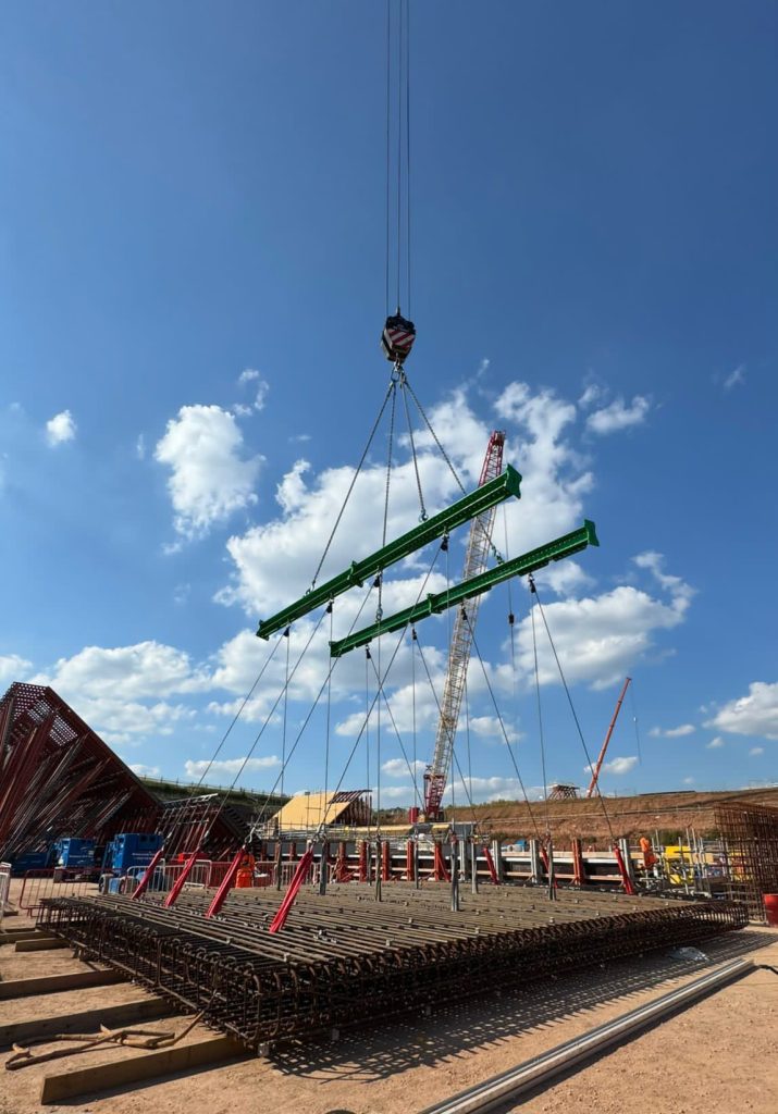 A construction site with a crane lifting a section of steel rebar against a clear blue sky, resembling an Auto Draft blueprint come to life. Other construction machinery and materials are visible in the background.