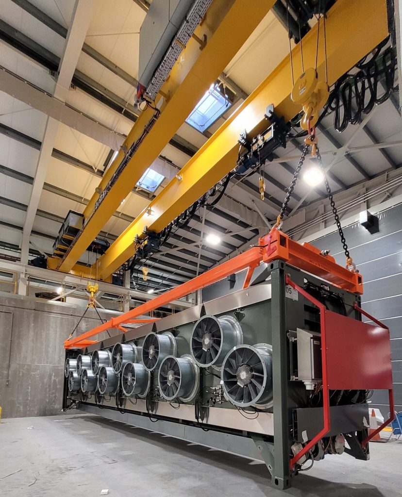 A large industrial fan unit, designed for direct air capture, is suspended from an overhead crane within a facility featuring a high ceiling and bespoke container frame.