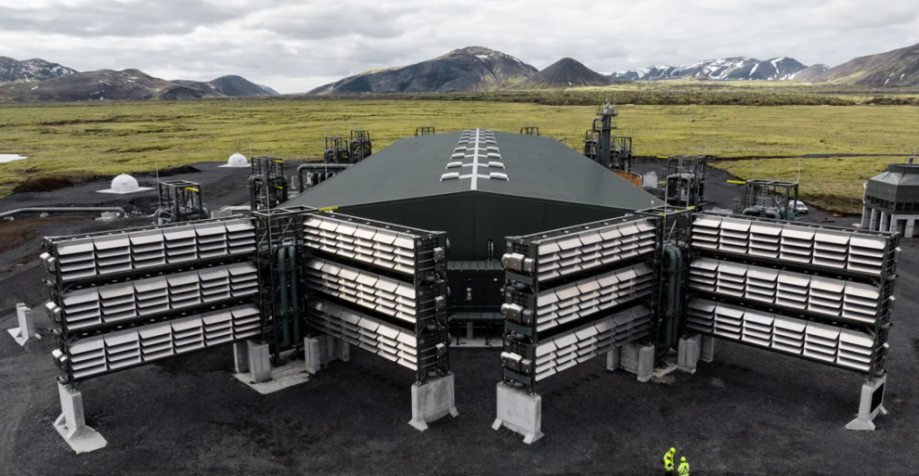 A large green industrial building, reminiscent of a pioneering facility, with vent structures nestled in a mountainous landscape; two people in safety gear stand nearby.