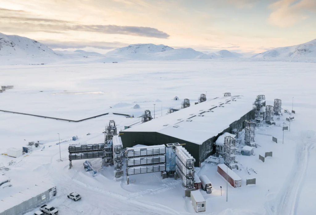 A large, industrial facility nestled in a snowy, mountainous landscape at dusk, features snow-covered roads and several stacks of equipment housed within container frames outside.