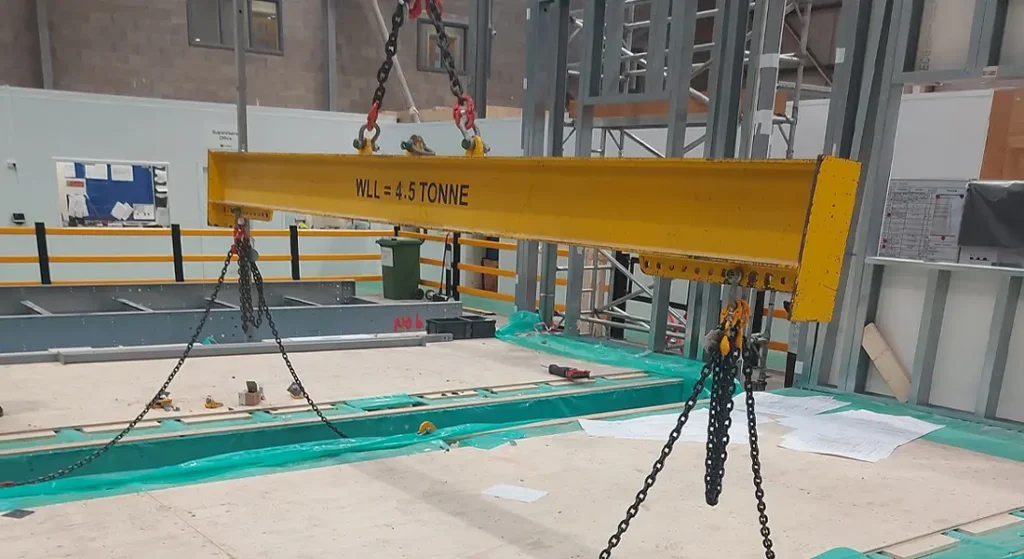 A yellow lifting beam labeled "WLL = 4.5 TONNE" is suspended by chains at a modular building construction site, with various materials and tools visible on the floor below, highlighting a focus on productivity. A yellow lifting beam labeled "WLL = 4.5 TONNE" is suspended by chains at a modular building construction site, with various materials and tools visible on the floor below, highlighting a focus on productivity.