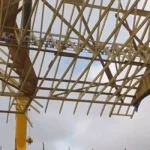 A construction crane, equipped with a Britlift modular roof truss lifter, hoists a large roof truss into position above a building under construction, with scaffolding surrounding the structure.