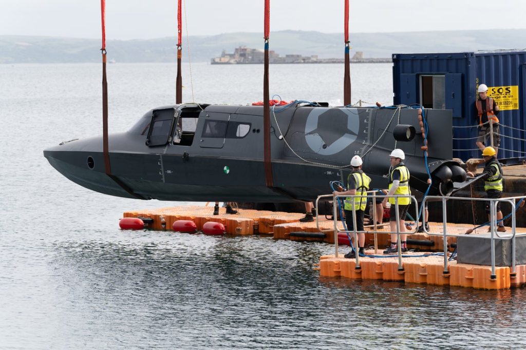 A black submersible is being lowered into the water from a quay using red Britlift straps, whilst workers in safety gear supervise this maritime defence operation. A black submersible is being lowered into the water from a quay using red Britlift straps, whilst workers in safety gear supervise this maritime defence operation.