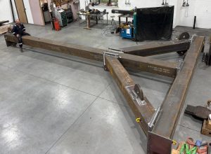 A large, welded steel structure with bespoke beams and triangular supports sits in a workshop. A person is seated on one beam, showing the scale of the metal assembly. Tools and equipment are visible nearby. A large, welded steel structure with bespoke beams and triangular supports sits in a workshop. A person is seated on one beam, showing the scale of the metal assembly. Tools and equipment are visible nearby.