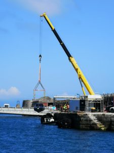 A yellow Britlift crane lifts a small boat out of the water onto a quay, with several workers—possibly part of a maritime defence team—standing nearby under a clear blue sky. A yellow Britlift crane lifts a small boat out of the water onto a quay, with several workers—possibly part of a maritime defence team—standing nearby under a clear blue sky.