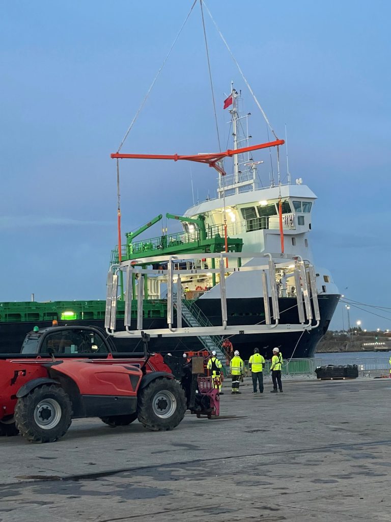 A group of workers in safety gear oversee a large metal structure, possibly bespoke beams from Britlift, being lifted by a crane near a docked cargo ship—an essential step for wind turbines installation. A group of workers in safety gear oversee a large metal structure, possibly bespoke beams from Britlift, being lifted by a crane near a docked cargo ship—an essential step for wind turbines installation.