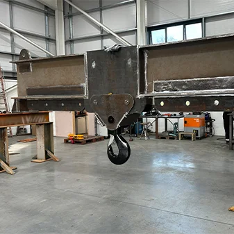 A large industrial crane hook and lifting beams attached to a steel beam structure inside a workshop, with various tools and equipment in the background.