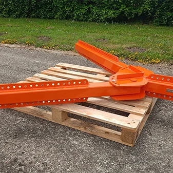 An orange metal lifting beam support structure with multiple holes rests on a wooden pallet outdoors on concrete, with grass and bushes in the background.
