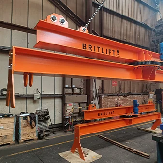 An orange lifting beam labelled "BRITLIFT" is suspended by a chain in an industrial warehouse, showcasing how lifting beams are used to safely handle heavy equipment and materials in demanding environments.