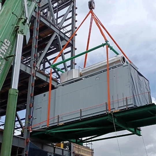 A large industrial unit is being lifted by specialised lifting equipment and orange straps at a construction site, with steel framework visible in the background.