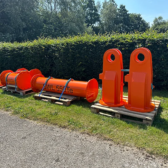 Four large orange metal components, likely parts of lifting equipment, including cylindrical and upright pieces, sit on wooden pallets beside a grassy area and hedge on a sunny day.