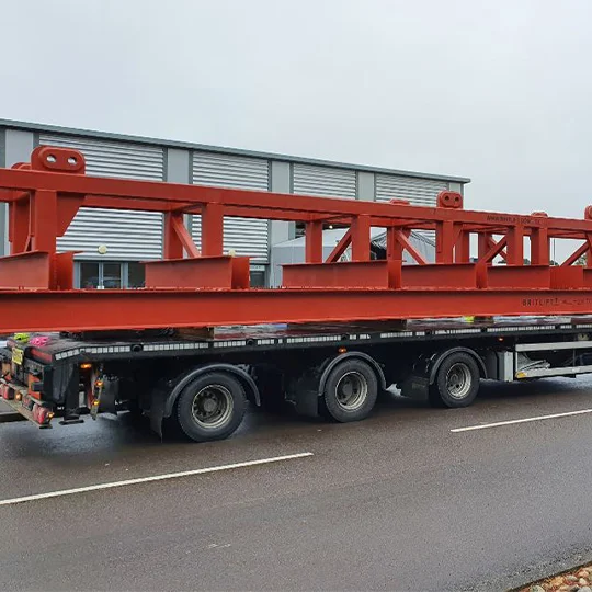 A large red metal framework, likely used as lifting equipment, is loaded onto a flatbed lorry parked on a road in front of an industrial building.