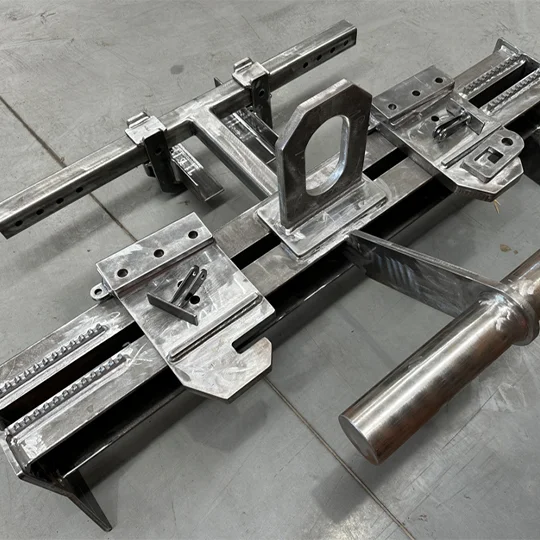 A metal welded fixture with brackets, plates, and cylindrical components, typically used as lifting equipment, arranged on a concrete floor.