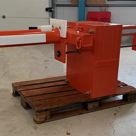 An orange mechanical barrier gate, often used with lifting equipment, is mounted on a wooden pallet and positioned indoors on a concrete floor.