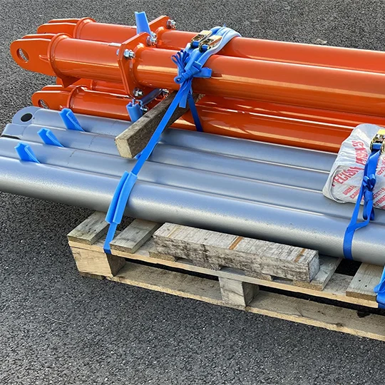 Several orange and grey metal pipes are strapped to a wooden pallet with blue ties, resting on a tarmac surface—ready for use with lifting equipment.