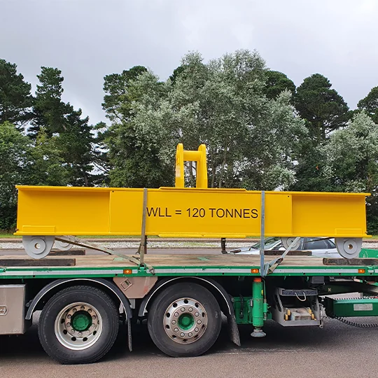 A large yellow metal beam labelled "WLL = 120 TONNES," part of heavy lifting equipment, is secured on a flatbed lorry, with trees visible in the background.