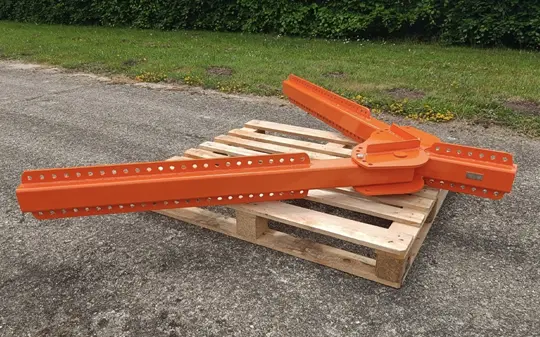 An orange metal beam structure, possibly part of bespoke lifting equipment, with multiple holes rests on a wooden pallet outdoors on a paved surface with grass and bushes in the background.